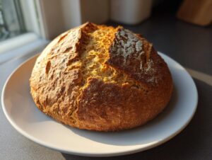 A golden brown, round loaf of freshly baked soda bread sitting on a white plate, highlighted by sunlight.
