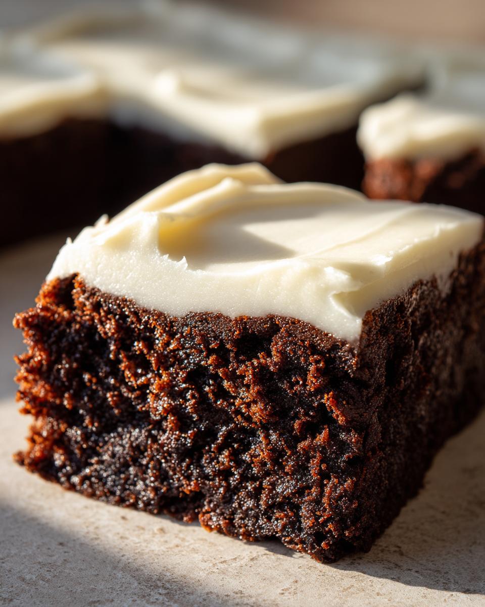 A close-up, macro shot of a rich, dark slice of gingerbread brownies topped with thick white cream cheese frosting.
