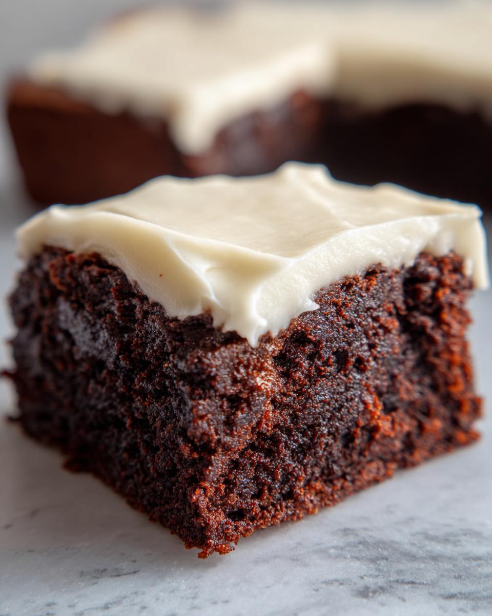 A close-up shot of a rich, dark square of gingerbread brownies topped with thick, creamy white frosting.