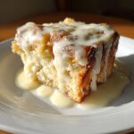 A close-up of a square slice of golden brown bread pudding generously topped with white vanilla sauce.