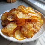 A close-up of a white bowl piled high with crispy, golden homemade potato chips sprinkled generously with salt.