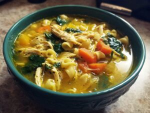 A close-up of a bowl of rich, yellow italian penicillin soup filled with shredded chicken, carrots, spinach, and pasta.