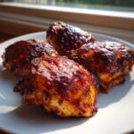 Four pieces of juicy bbq chicken, heavily glazed with dark, sticky sauce, resting on a white plate.