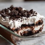 A rich, layered dirt cake showing chocolate pudding, cream filling, and crushed Oreo topping in a clear glass baking dish.