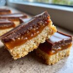 Two stacked squares of homemade Twix bars showing the shortbread base, gooey caramel, and chocolate topping.