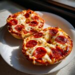 Two halves of freshly baked pizza bagels topped with melted mozzarella, pepperoni slices, and herbs on a white plate.