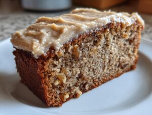 Close-up of a moist slice of applesauce cake topped with thick, creamy brown sugar frosting on a white plate.