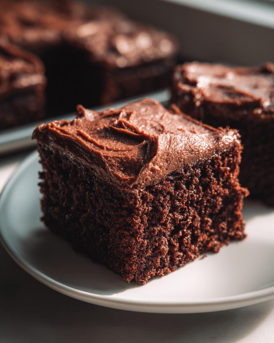 Close-up of a square slice of dark, moist gingerbread cake topped with thick chocolate frosting on a white plate.