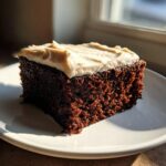 A close-up, sunlit slice of dark, moist gingerbread cake topped with thick, light brown frosting.