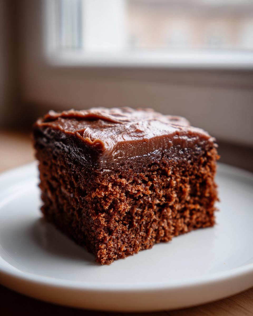 A close-up of a single, moist square slice of dark gingerbread cake topped with rich chocolate frosting.