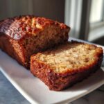 Close-up of a moist greek yogurt banana bread loaf, with one thick slice cut and resting beside it.