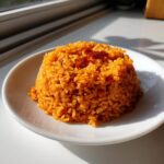 A molded mound of vibrant orange jollof rice served on a small white plate, highlighted by natural sunlight.