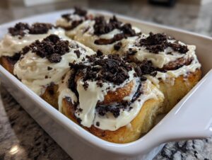 A close-up of freshly baked Oreo cinnamon rolls topped with thick white frosting and crushed chocolate cookies.