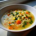 A comforting bowl of penicillin soup featuring small round pasta, diced carrots, and fresh parsley garnish in a clear broth.