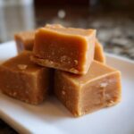 A stack of four golden-brown, creamy squares of homemade penuche fudge resting on a white plate.