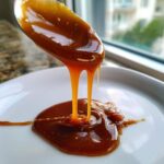 Close-up of thick, glossy homemade caramel sauce dripping from a spoon onto a white plate.