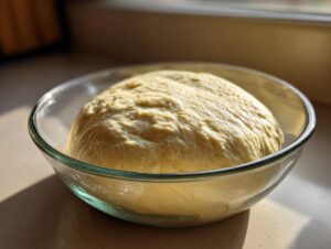 A large, smooth ball of risen pizza dough recipe resting in a clear glass bowl, catching warm sunlight.