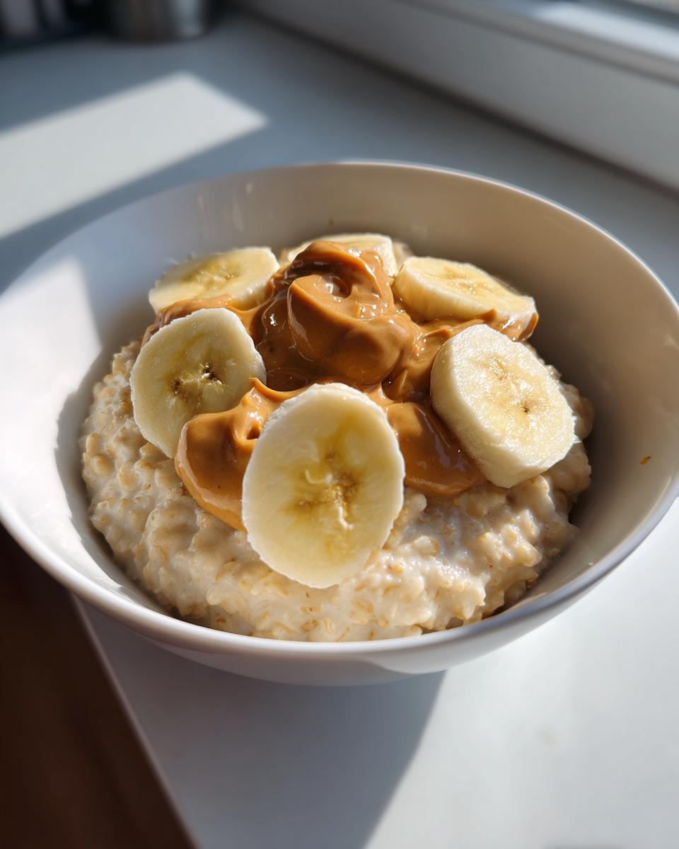 A white bowl filled with creamy banana oatmeal, topped with sliced bananas and a generous swirl of peanut butter.