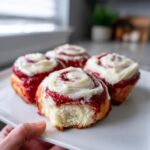 Four decadent red velvet cinnamon rolls topped with cream cheese frosting on a white plate, one has a bite taken out.