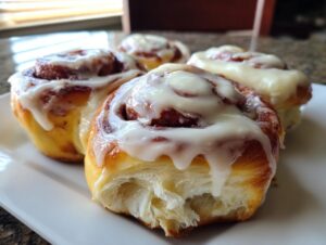 Four freshly baked red velvet cinnamon rolls generously topped with white cream cheese icing, featuring a soft, torn interior.