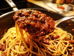 A spoonful of rich, thick bolognese sauce being lifted from a pan of spaghetti noodles.
