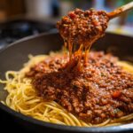 A wooden spoon dripping rich, thick bolognese sauce over a bed of spaghetti noodles in a dark pan.