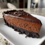 A decadent slice of chocolate cheesecake featuring a dark cookie crust, thick mousse filling, and glossy ganache topping.