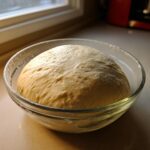 A large, risen ball of pizza dough recipe resting in a clear glass bowl near a window.