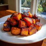 A white plate piled high with caramelized, dark-edged roasted sweet potatoes cubes, catching bright sunlight.