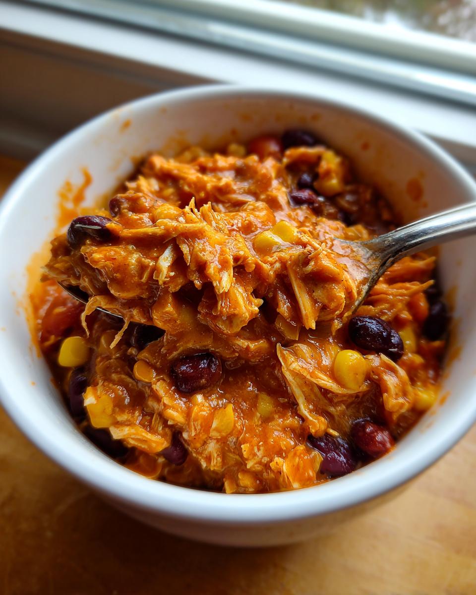 A spoonful of shredded slow cooker buffalo chicken chili, mixed with kidney beans and corn, being lifted from a white bowl.