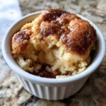 A close-up view of a warm snickerdoodle cobbler dessert served in a white fluted ramekin, showing a gooey cinnamon topping.
