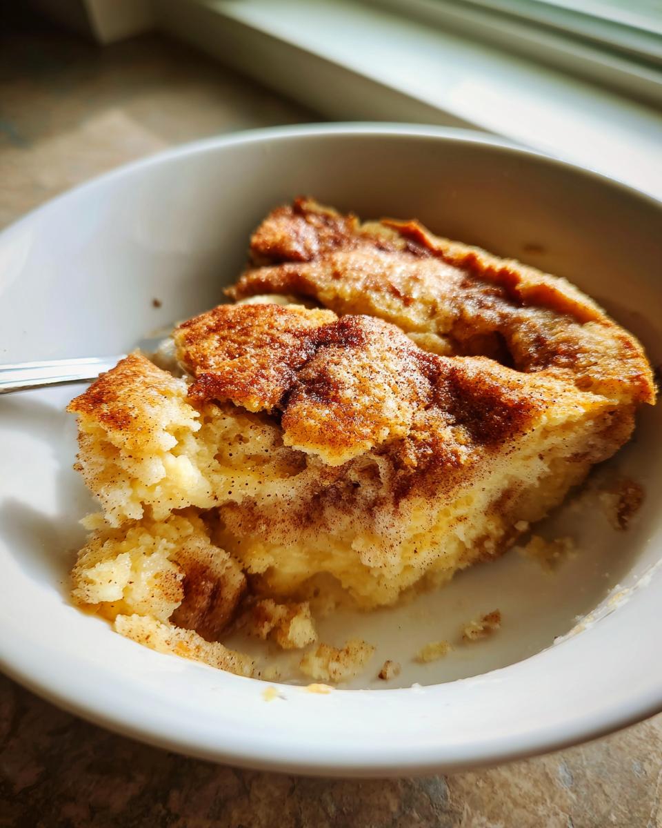A close-up of a warm serving of snickerdoodle cobbler, topped with cinnamon sugar, inside a white bowl.