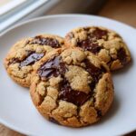 Close-up of three freshly baked sourdough chocolate chip cookies with melted chocolate chunks on a white plate.