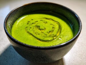Close-up of a vibrant green zucchini soup, drizzled with olive oil and pepper, served in a dark ceramic bowl.