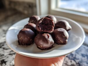 A hand holds a white plate piled with chocolate-dipped Oreo balls, one of which is broken open to show the dark interior.
