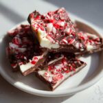 A stack of rich, layered peppermint bark pieces topped with crushed candy canes on a white plate.