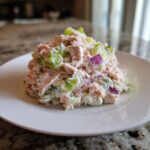 A mound of creamy salmon salad mixed with celery, lettuce, and red onion served on a white plate.