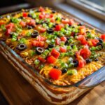 Close-up of an amazing 7 layer dip topped with melted cheese, diced tomatoes, olives, and green onions.