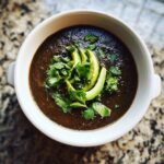 Overhead view of a bowl of rich, dark black bean soup garnished with sliced avocado and fresh cilantro.