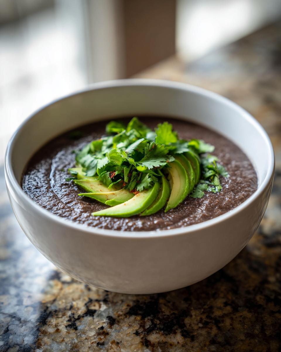 A close-up of a bowl of creamy black bean soup garnished with sliced avocado and fresh cilantro.