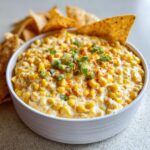 A creamy, cheesy corn dip topped with green onions and paprika, served with tortilla chips for dipping.