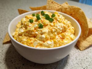 A creamy, bright yellow corn dip topped with green onions and paprika, served with tortilla chips for dipping.