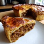 A thick slice of amazing meat pie with a rich, savory filling and golden brown crust, served on a white plate.
