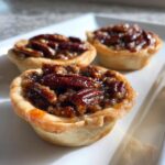 Three glistening mini pecan pies with flaky crusts and caramelized pecan toppings, ready to eat.