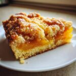 Close-up of a single serving slice of peach cobbler showing a thick layer of orange fruit filling and a golden crumb topping.