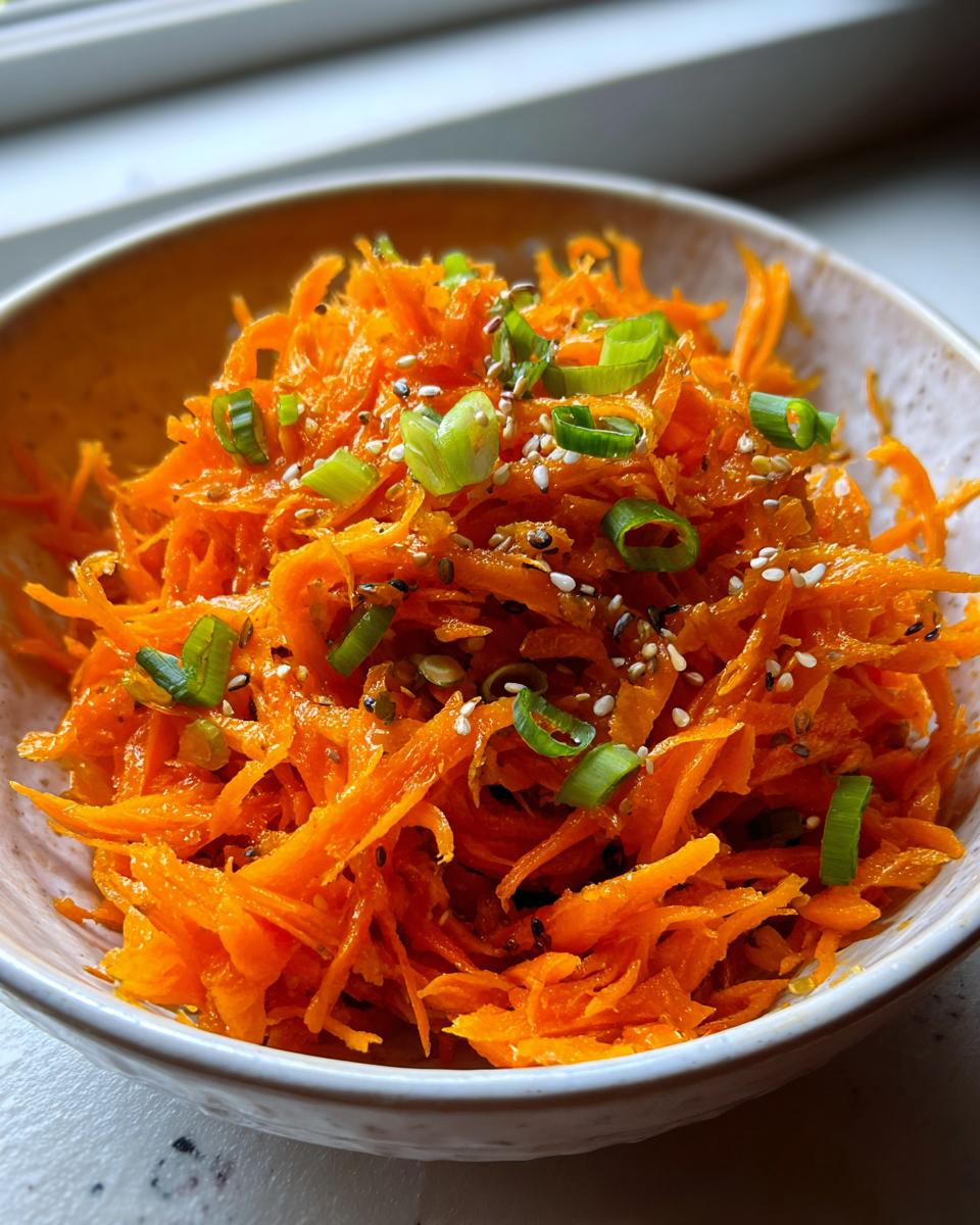A close-up of a vibrant orange raw carrot salad, shredded and topped with green onions and sesame seeds.
