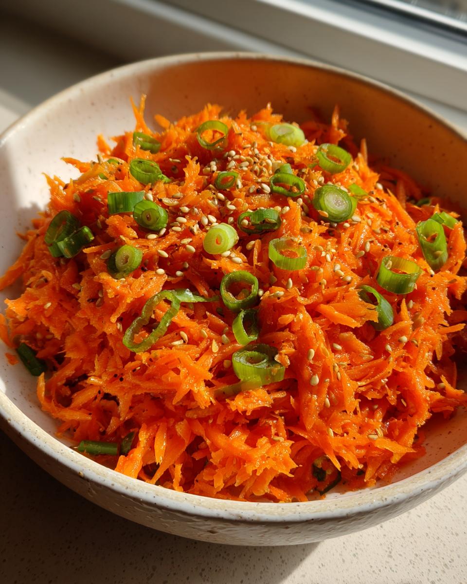 Close-up of a vibrant raw carrot salad, shredded and topped with sliced green onions and sesame seeds.
