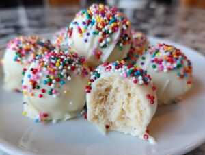 Close-up of several white chocolate-coated sugar cookie truffles topped with colorful sprinkles, one is bitten open.