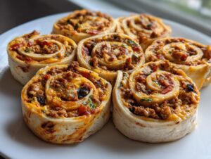Close-up of six baked taco pinwheels filled with seasoned ground meat, cheese, and vegetables, arranged on a white plate.
