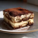 A perfect square slice of rich tiramisu recipe layered with cream and ladyfingers, dusted heavily with cocoa powder.
